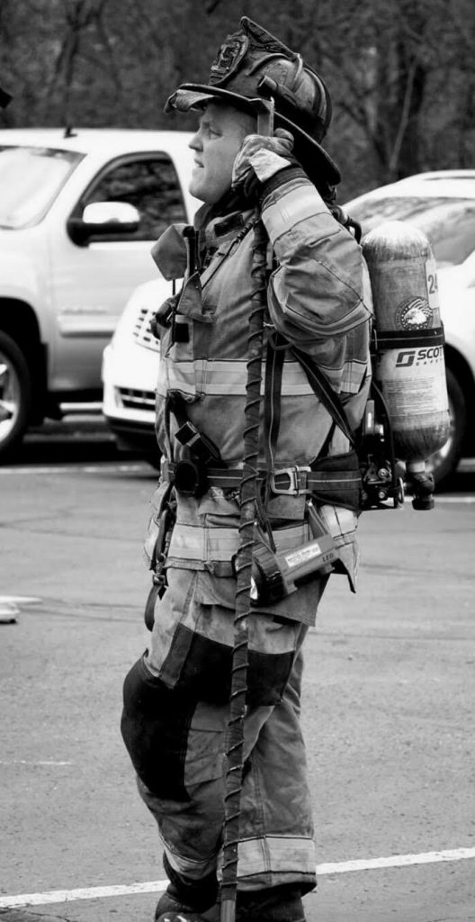 A firefighter in full gear and helmet, equipped with an oxygen tank, stands in a parking lot. He is holding a communication device to his ear, appearing to be listening closely. Parked vehicles are visible in the background. The image is in black and white.
