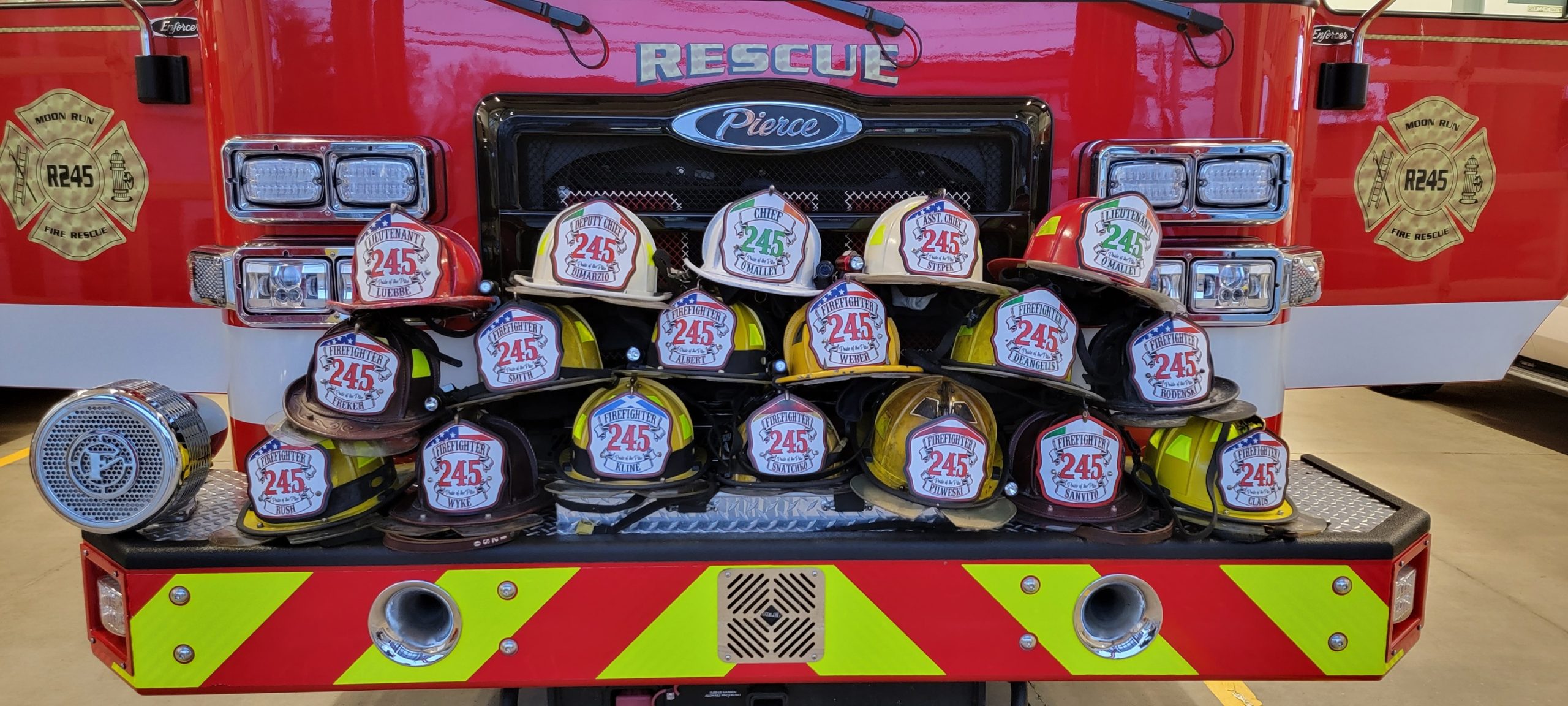 A display of various firefighter helmets, each with "245" on the front, arranged on the front bumper of a red rescue fire truck parked in a station. Two fire department logos are visible on either side of the truck.
