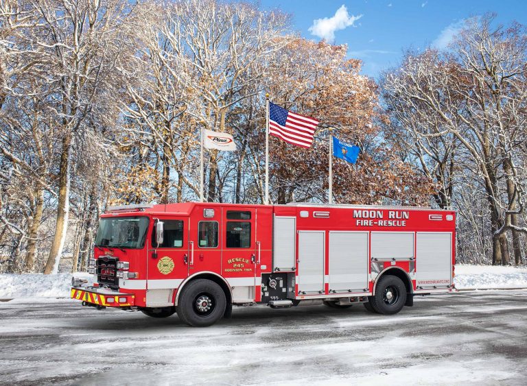 A bright red fire-rescue truck labeled "Moon Run" is parked on a snow-covered road. The truck is positioned against a backdrop of snowy trees and a clear blue sky. Three flags (American, state, and another) are displayed on poles behind the truck.