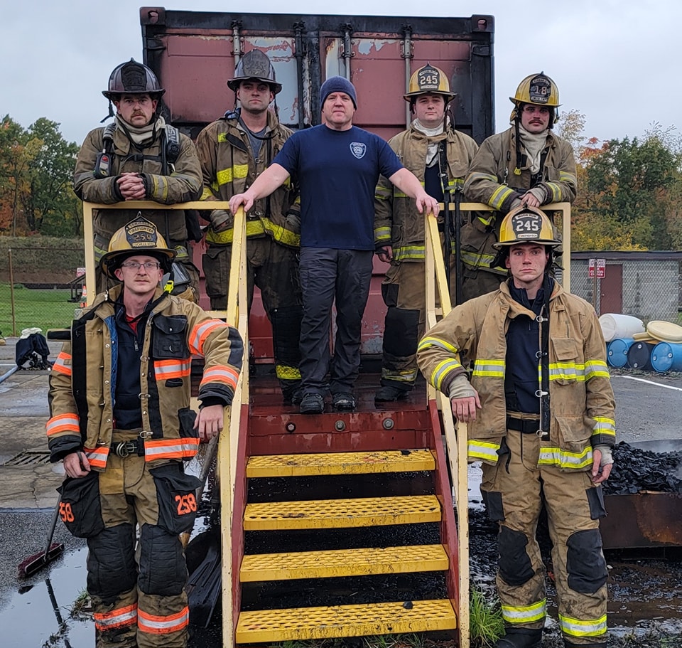 A group of six firefighters in gear stands together on a set of yellow stairs attached to a firefighting training structure. Some wear helmets with numbers, and the central figure at the top of the stairs is in a blue shirt and beanie. The background is cloudy.