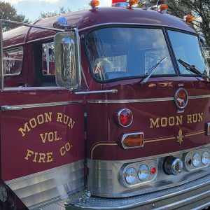 A vintage fire truck from Moon Run Volunteer Fire Company with an open driver's side door. The truck is painted in deep red and features chrome accents, red emergency lights, and the words "Moon Run Vol. Fire Co." emblazoned on the side and front.